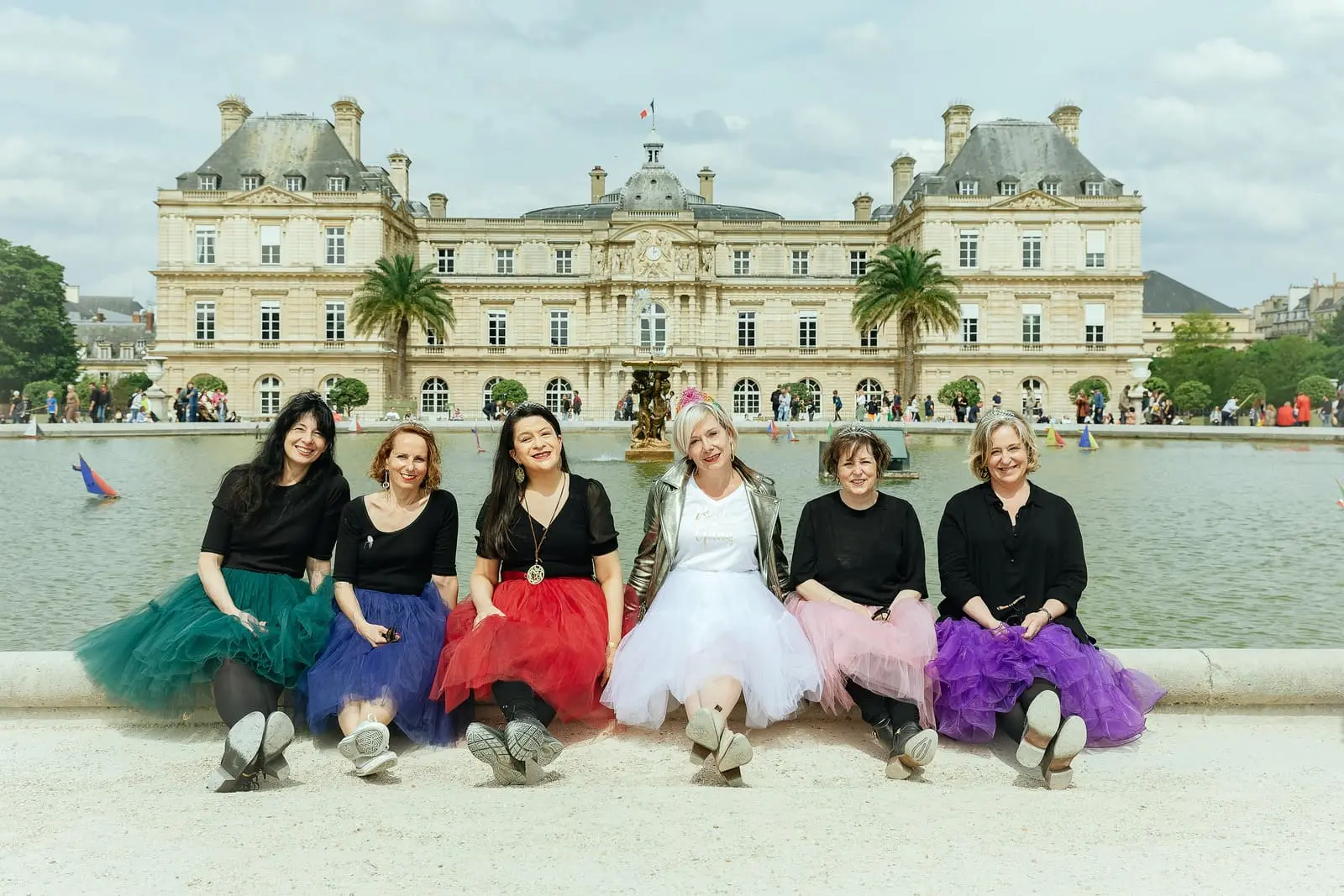 Un groupe de filles prend la pose, assises, au bord de la grande Fontaine du Jardin du Luxembourg, dos au Palais, lors d'une séance photo EVJF.