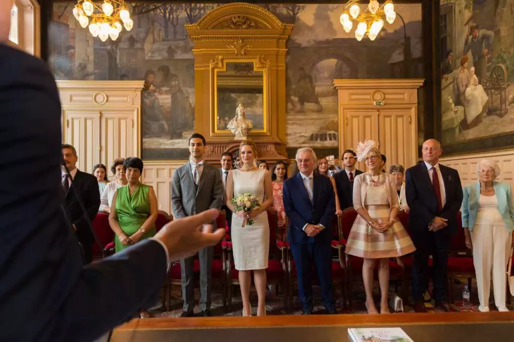 Photographie d'un mariage civil à la mairie de Paris 15 : la mariée, en tailleur, porte un bouquet de fleurs et se tient à proximité de son époux